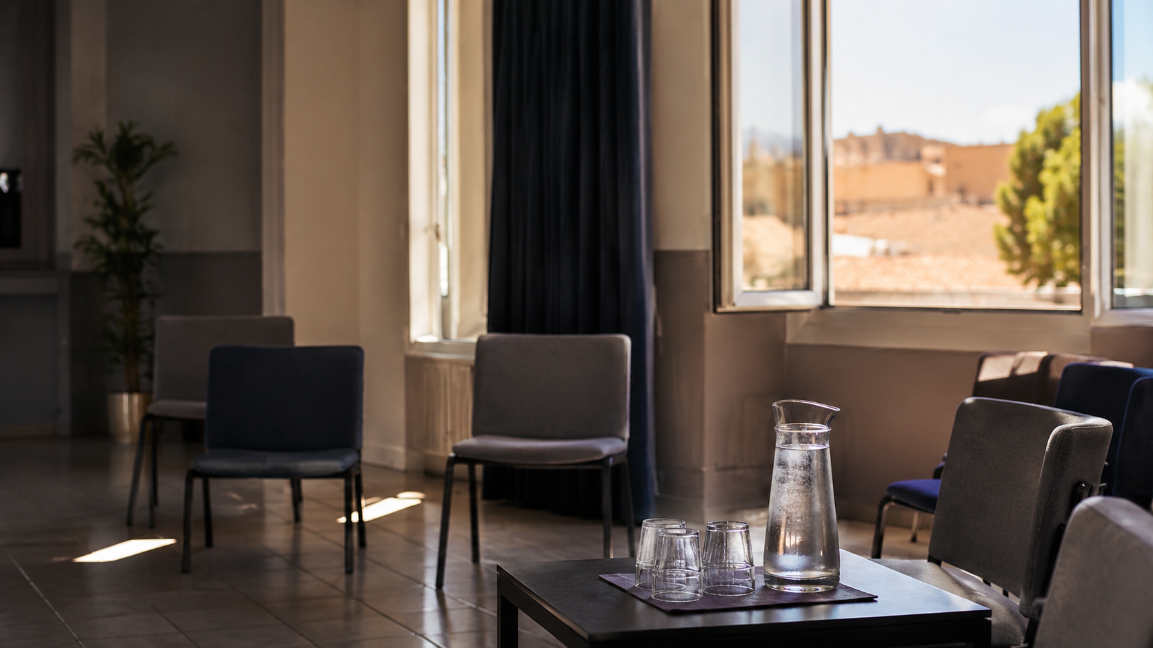 Une salle municipale fraîche avec chaises, carafe d’eau et rideaux tirés, sans personne identifiable.