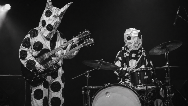 Deux musiciens masqués en costumes à pois noir et blanc sur une scène nue, guitare double manche et batterie