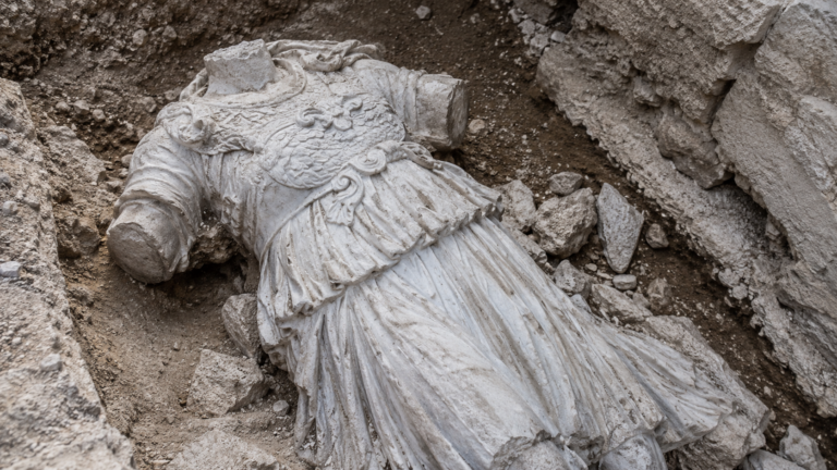 Statue de marbre blanc drapée, sans tête ni bras, couchée parmi des pierres sur un site de fouille