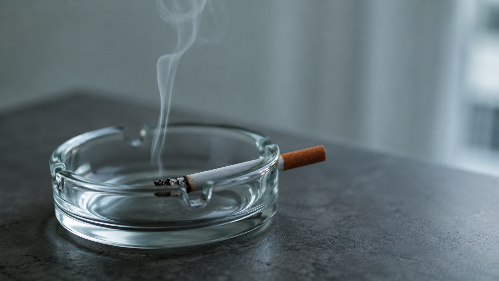 Une cigarette posée dans un cendrier propre, un mince filet de fumée, lumière froide