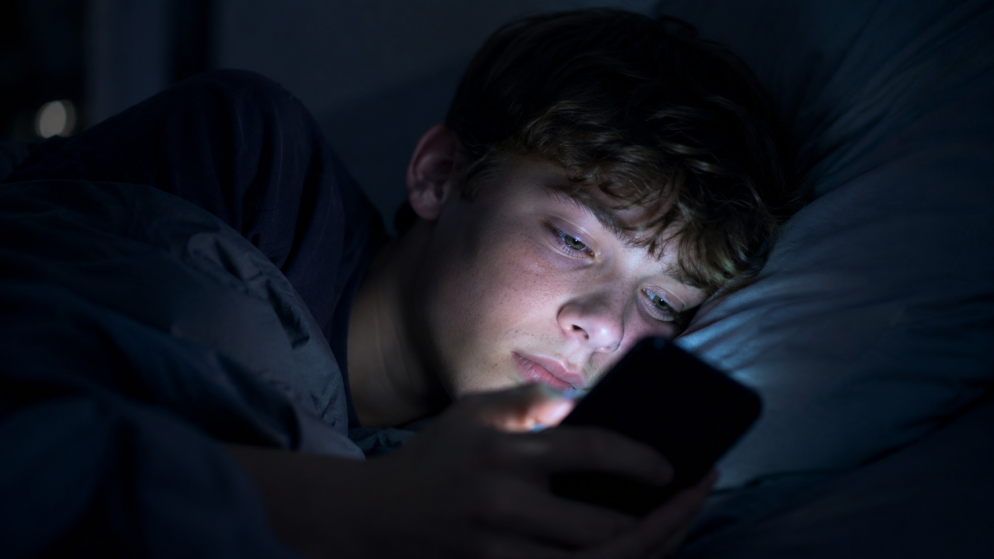 Adolescent allonge dans le noir, visage eclaire par l'ecran d'un telephone, regard en alerte