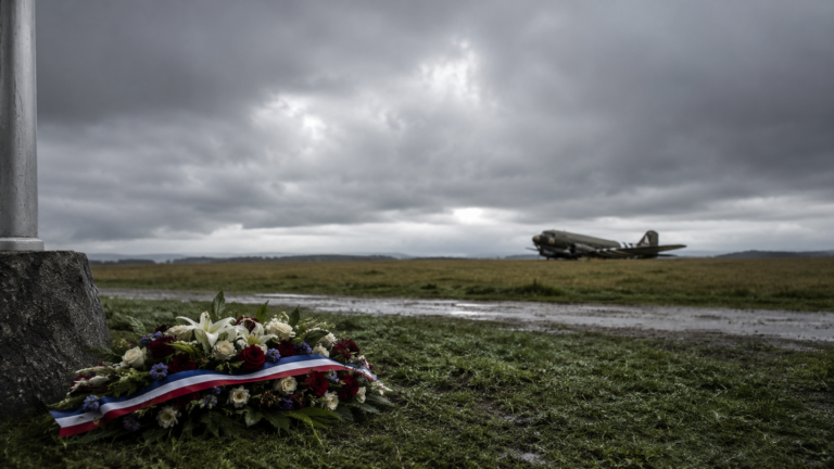 Un champ normand sous ciel bas avec une gerbe de commémoration au premier plan.