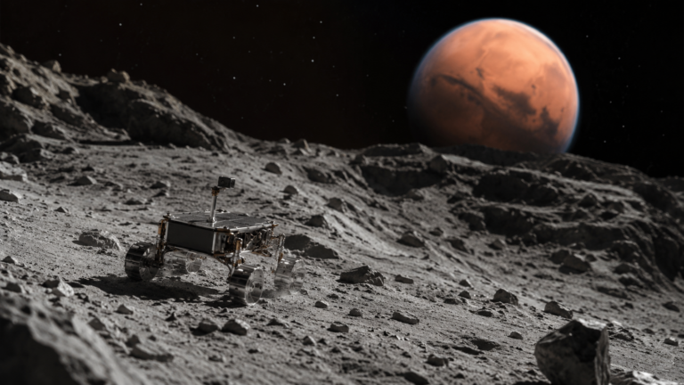 Petit rover spatial non marqué posé sur un sol gris évoquant Phobos, avec Mars floue au loin.