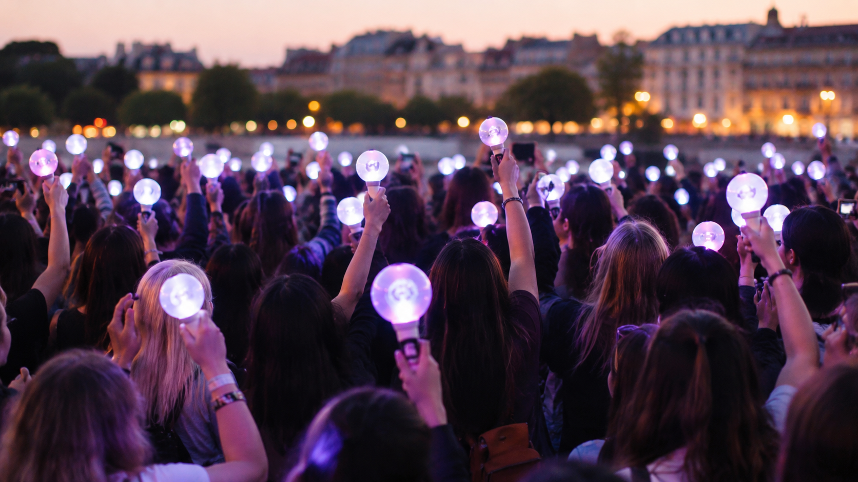 Foule de jeunes fans de K-pop vus de dos avec des light sticks lors d'un evenement en soiree