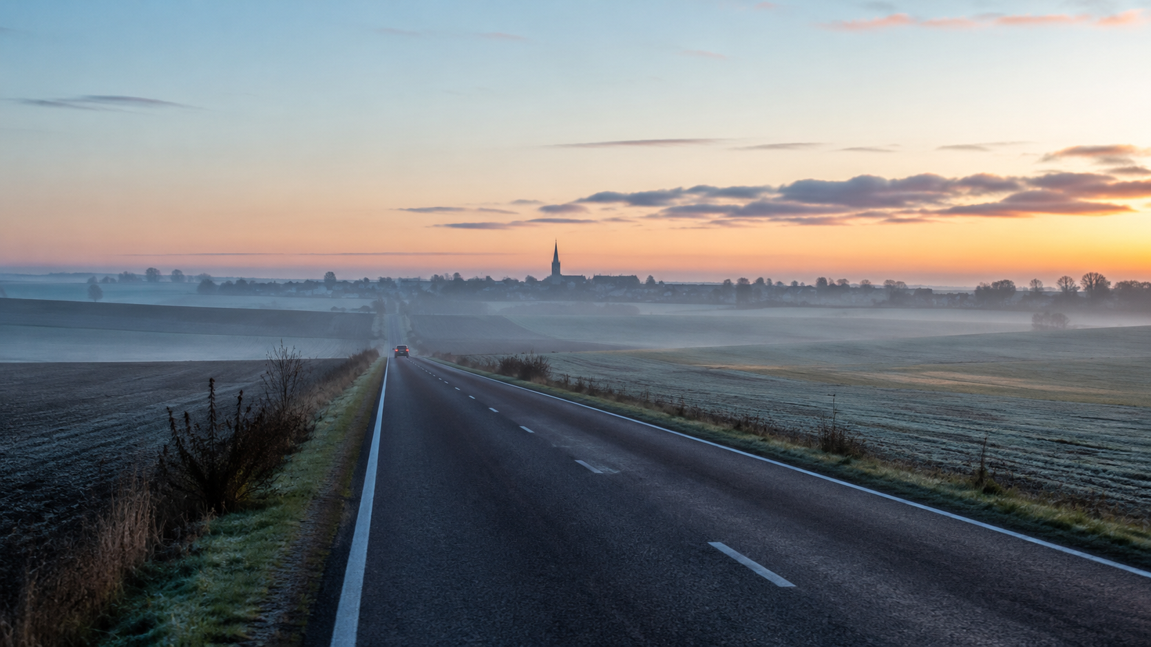 Une route de campagne à l'aube menant vers un village, une voiture au loin