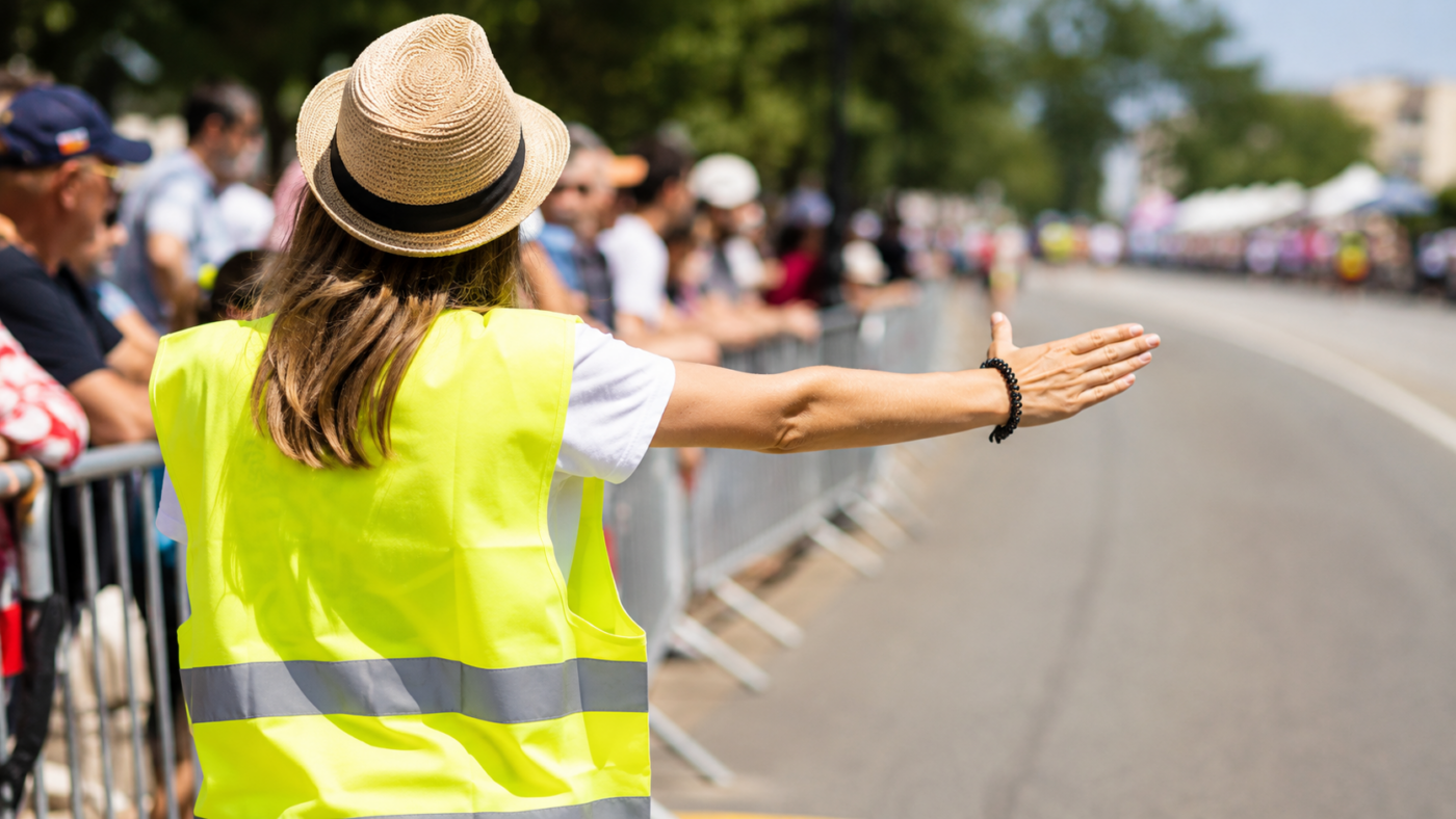 Bénévole en chasuble fluo orientant le public au bord d'une route avant une course cycliste