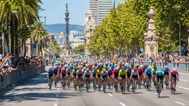 Un peloton de cyclistes traverse une grande avenue urbaine ensoleillee bordee de foule