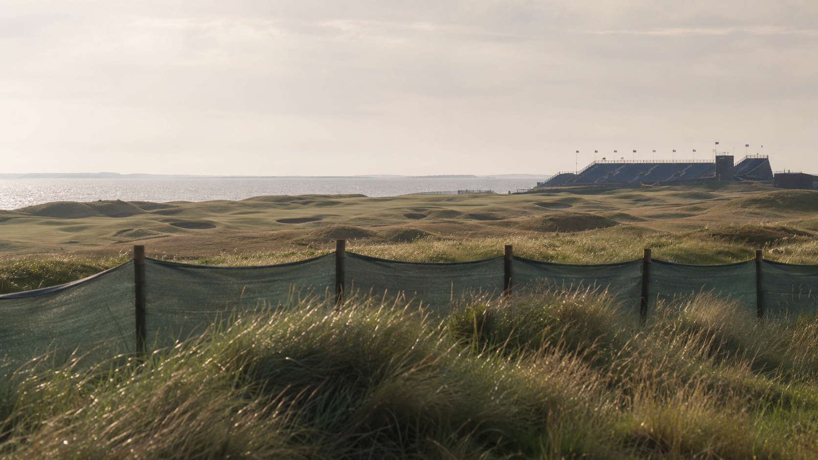 Un parcours de golf en bord de mer aperçu au loin derrière une clôture temporaire et des herbes de dune