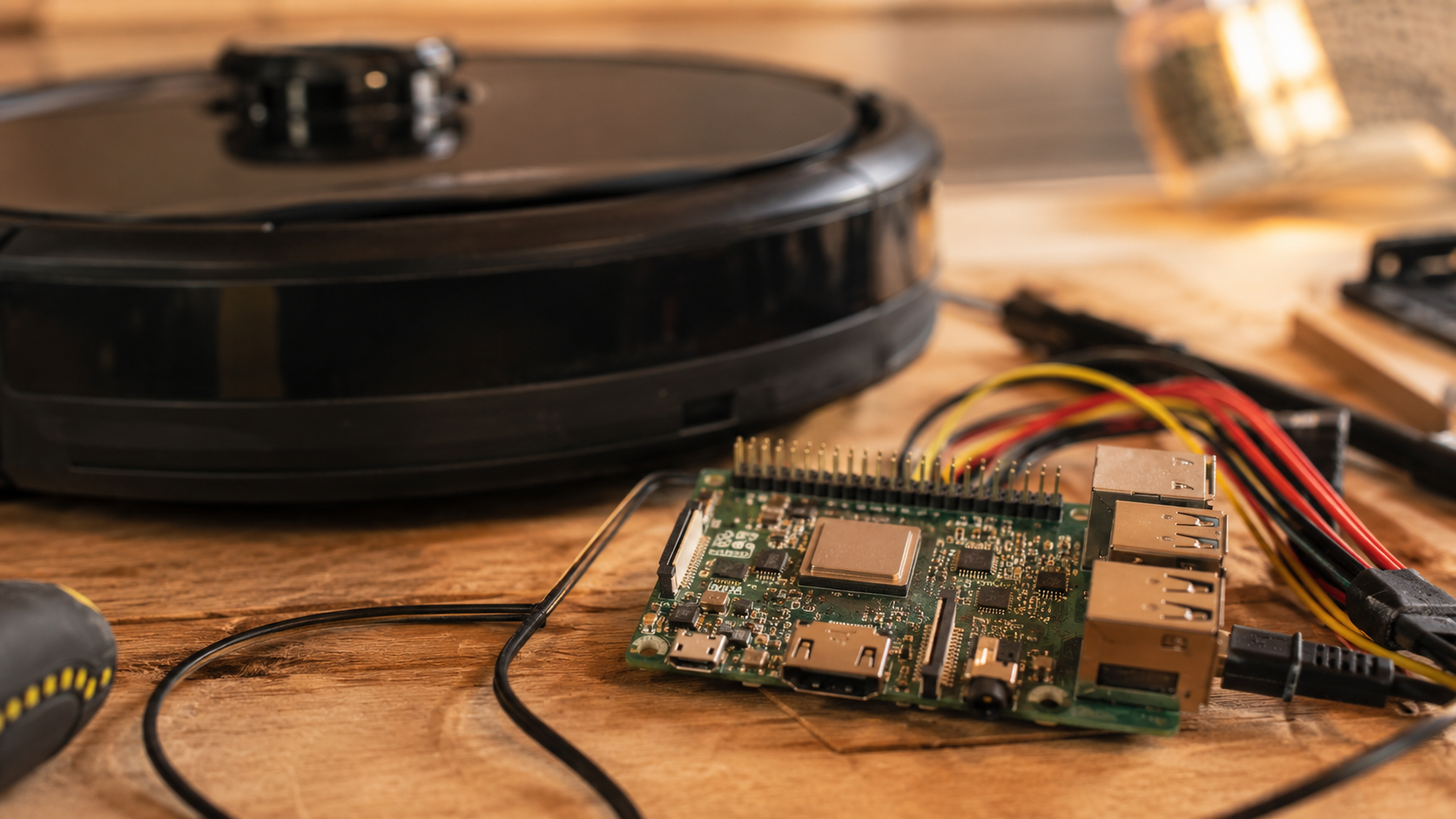 Une petite carte type Raspberry Pi et des cables sur une table en bois, un aspirateur robot flou a cote