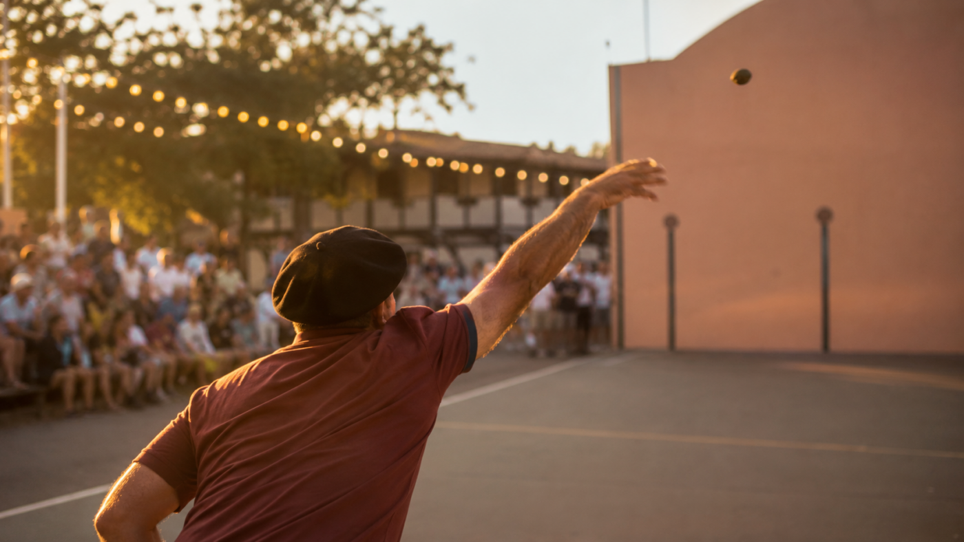 Personne anonyme vue de trois-quarts dos lançant une pigne de pin sur un fronton de pelote lors d'une fête d'été