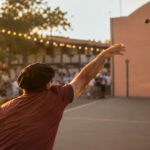 Personne anonyme vue de trois-quarts dos lançant une pigne de pin sur un fronton de pelote lors d'une fête d'été
