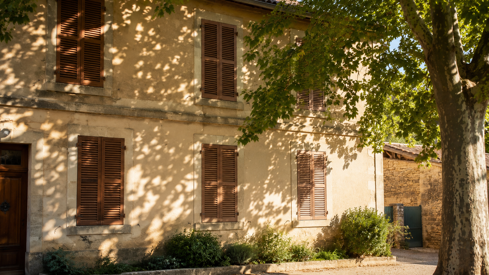 Une façade de maison aux volets de bois fermés dans la chaleur, un arbre jetant son ombre sur le devant