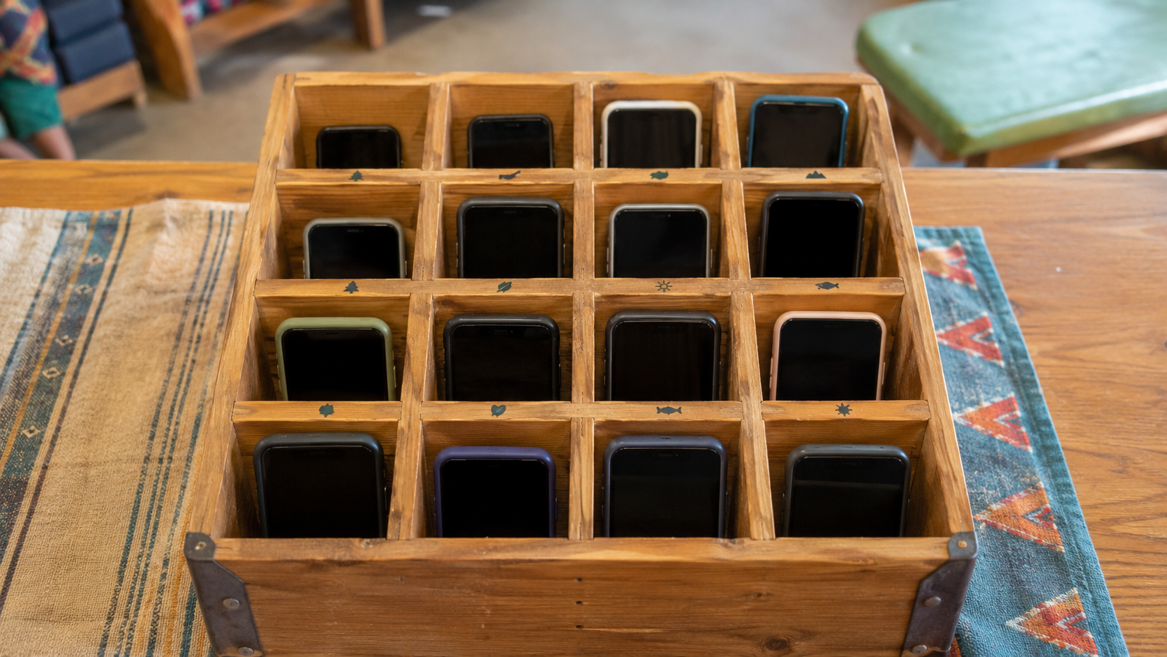 Une boîte en bois à casiers numérotés contenant plusieurs smartphones éteints, posée sur une table en colonie