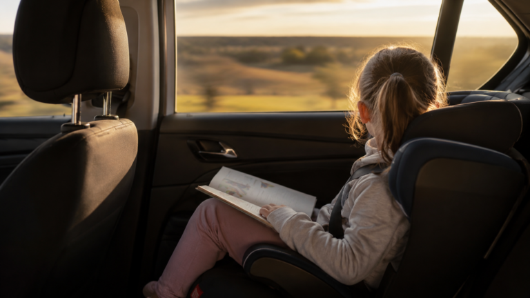 Jeune enfant vu de dos dans un siège auto, regardant par la vitre pendant un trajet d'été, un livre sur les genoux