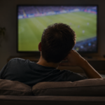 Une personne vue de dos sur un canapé, regardant seule un match de football à la télévision dans le noir