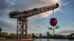 Grue de chantier historique a fleche cantilever au bord d'un fleuve, silhouette florale abstraite suspendue a son bras sous un ciel changeant