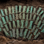 Une quarantaine de haches plates en bronze ancien posées ensemble sur une terre sombre au fond d'une fouille, patine vert-de-gris