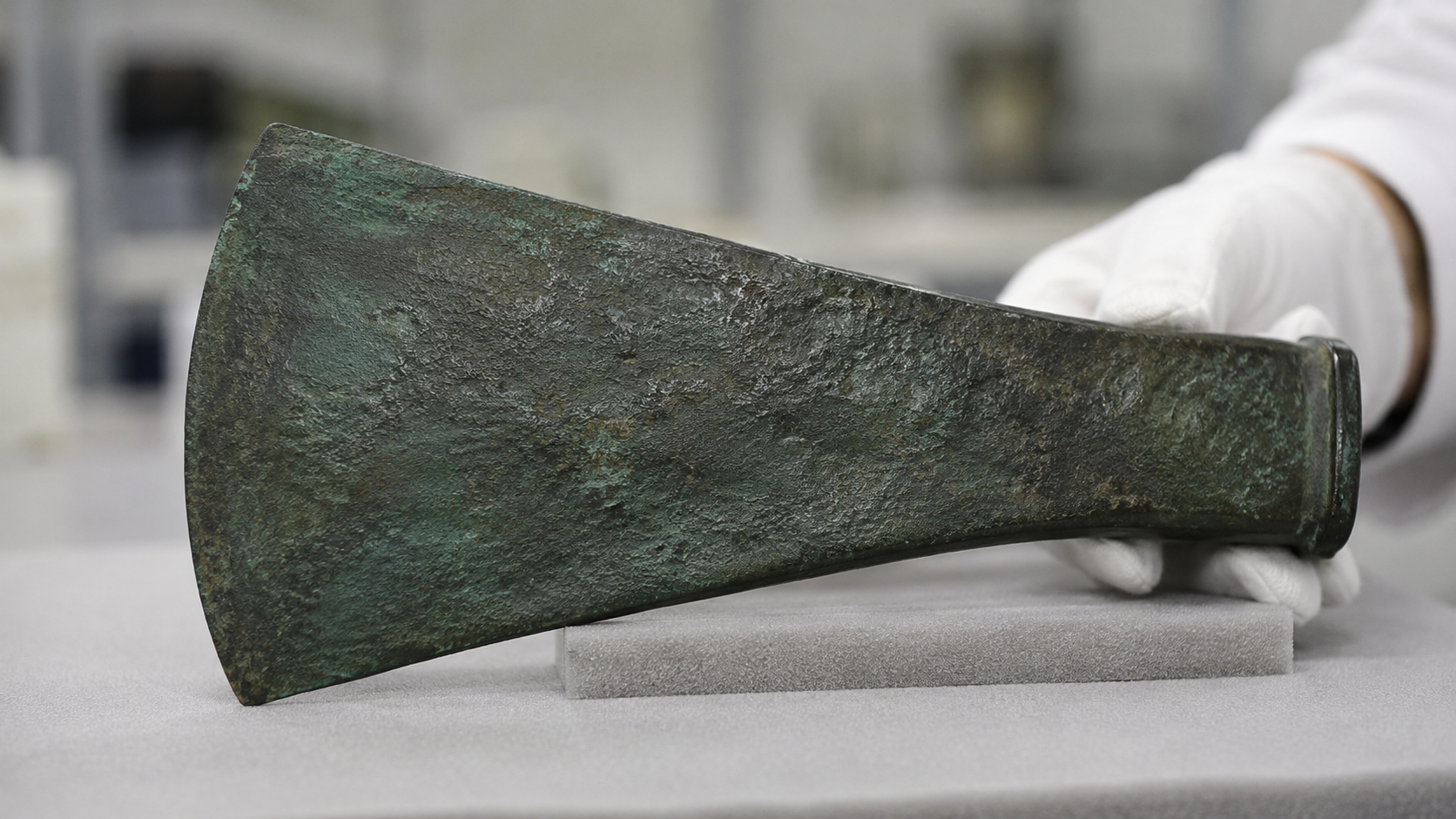 Une hache plate en bronze ancien tenue par une main gantée, lame coulée d'une pièce sans douille, patine vert-de-gris