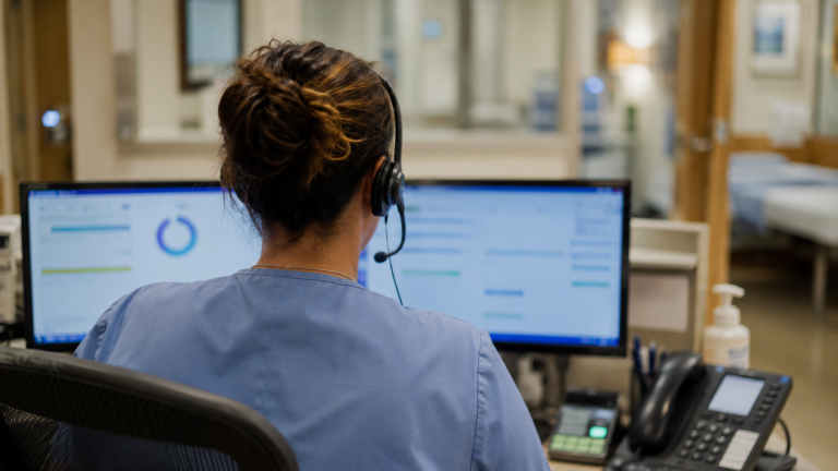 Une infirmière vue de dos à un poste d'appels, casque sur les oreilles, face à deux écrans affichant des graphiques et un minuteur