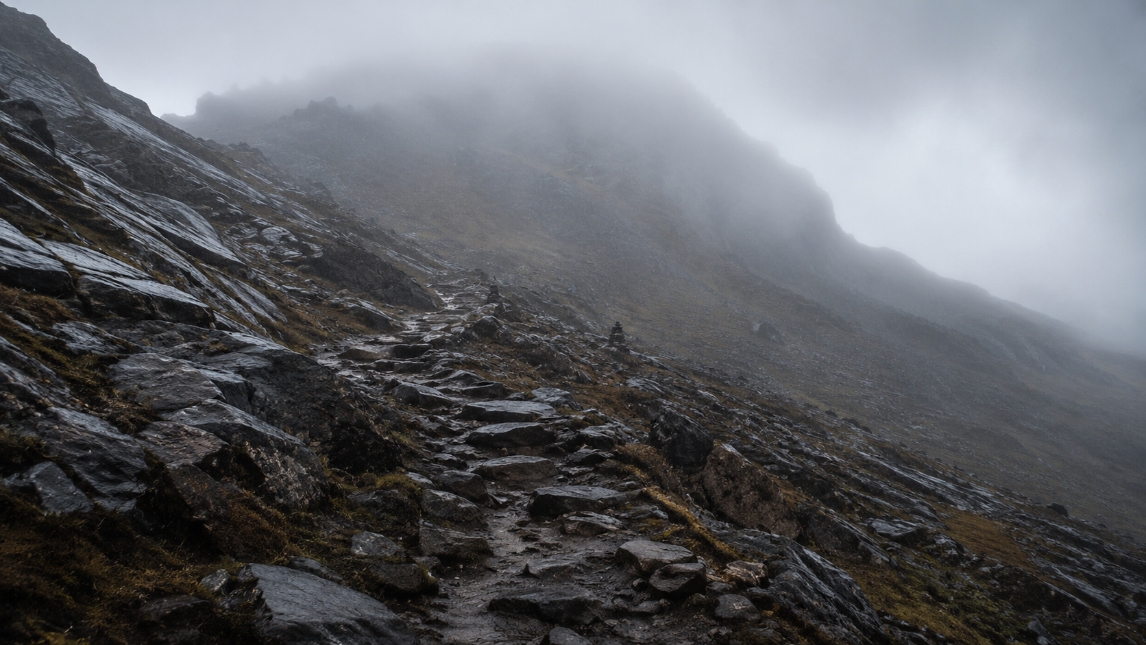 Un sentier de montagne caillouteux et raide qui disparaît dans des nuages bas, sous une pluie fine