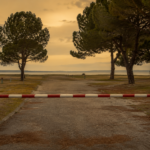 Barrière abaissée à l'entrée d'un camping littoral vide, emplacements herbeux déserts et pins maritimes sous un ciel ocre