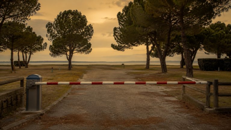 Barrière abaissée à l'entrée d'un camping littoral vide, emplacements herbeux déserts et pins maritimes sous un ciel ocre