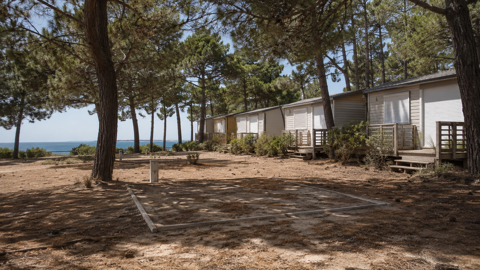 Rangée de mobil-homes aux volets fermés sur un camping sablonneux sous les pins, emplacement de tente vide au premier plan