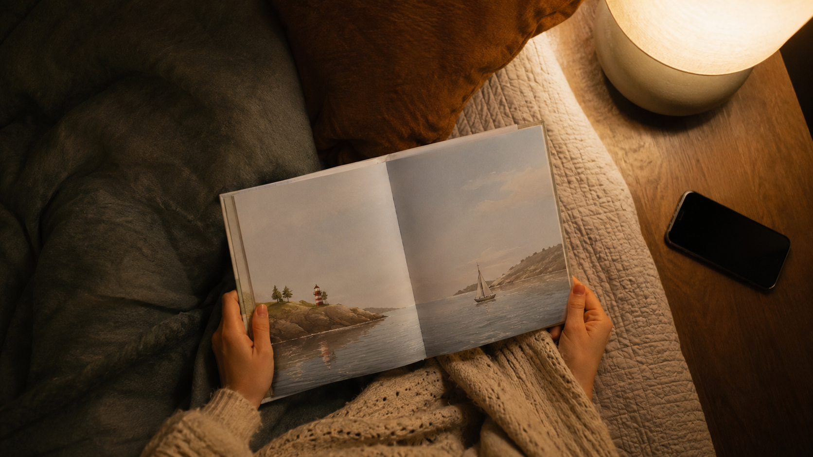 Mains d'adulte tenant un livre illustré ouvert sur un lit, un téléphone éteint posé de côté, vue de dessus