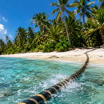 Câble à fibre optique sous-marin sortant d'une eau turquoise et remontant sur une plage de sable corallien bordée de cocotiers