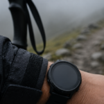 Gros plan d'un poignet portant une montre noire sans marque, main posée sur un bâton de randonnée, sentier rocheux et brume en arrière-plan