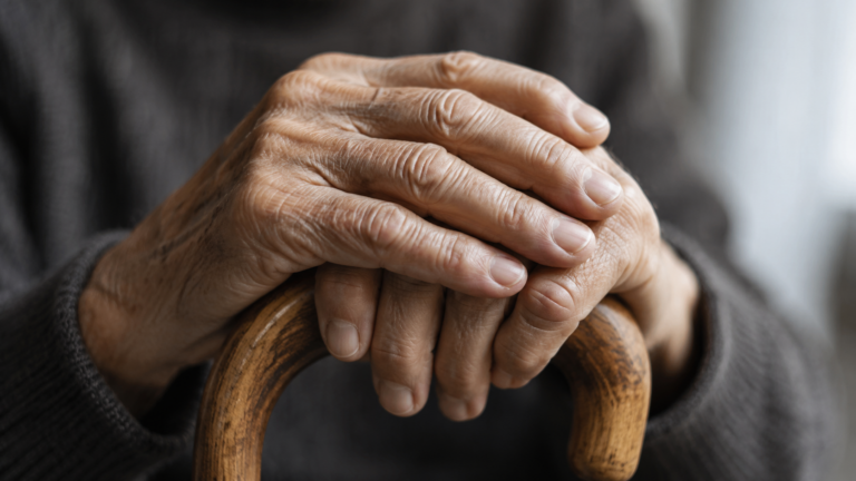Les mains agees d'une personne posees sur le pommeau d'une canne en bois, dans une lumiere douce