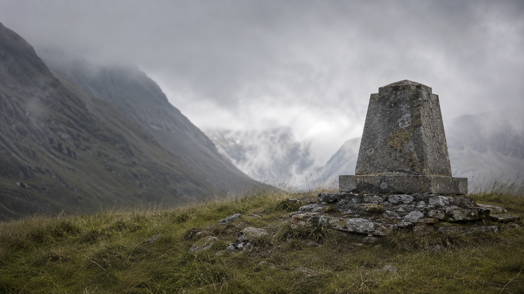 Un monument de pierre usé par le temps au sommet d'un col, dans l'herbe rase et les nuages bas