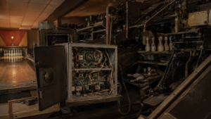 Intérieur d'un petit bowling ancien avec un coffret ouvert montrant câbles, cartes électroniques et relais reliés à la mécanique