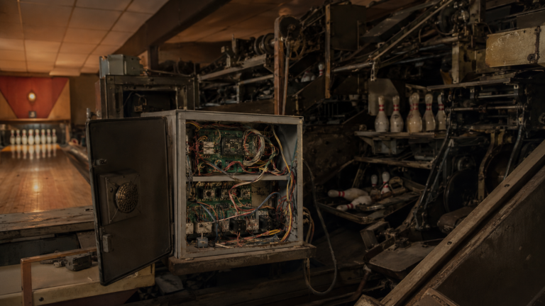 Intérieur d'un petit bowling ancien avec un coffret ouvert montrant câbles, cartes électroniques et relais reliés à la mécanique