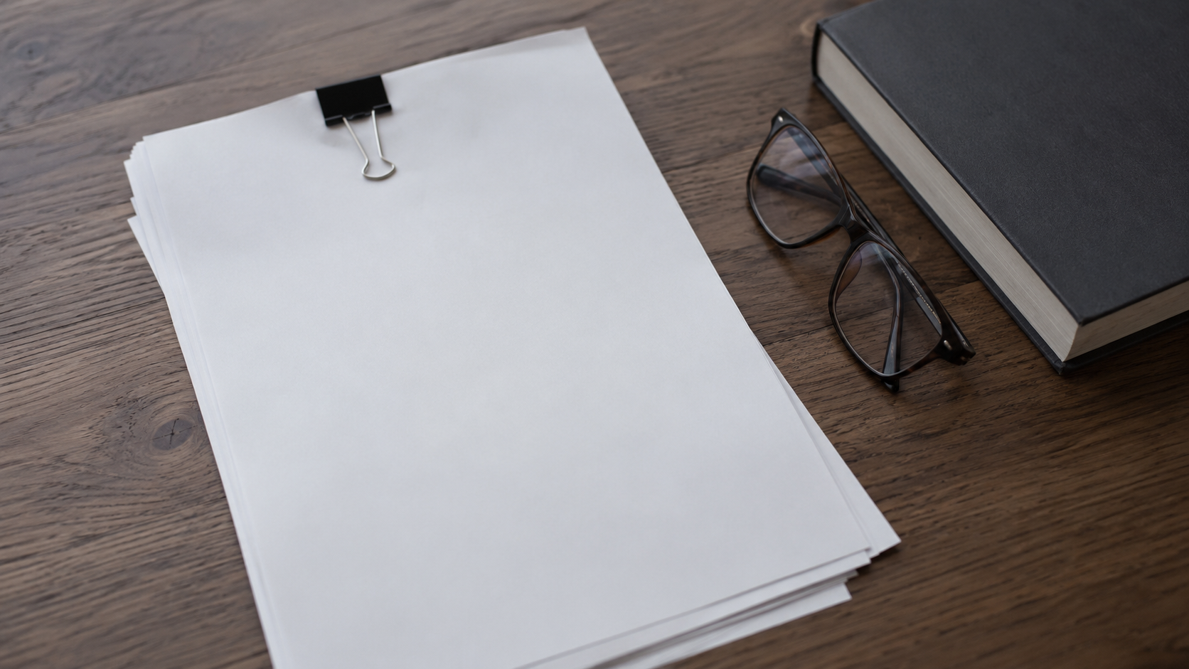 Une pile de contrats vierges tenue par une pince sur un bureau en bois, à côté d'une paire de lunettes