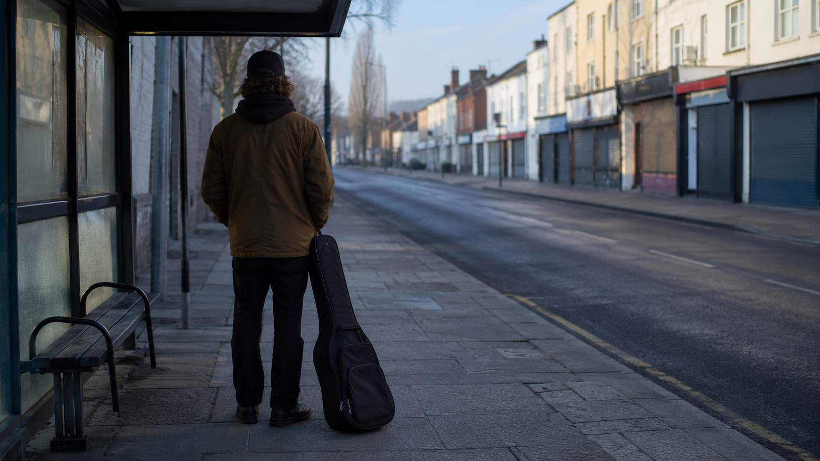Un musicien vu de dos à un arrêt de bus au petit matin, sa housse d'instrument posée contre sa jambe