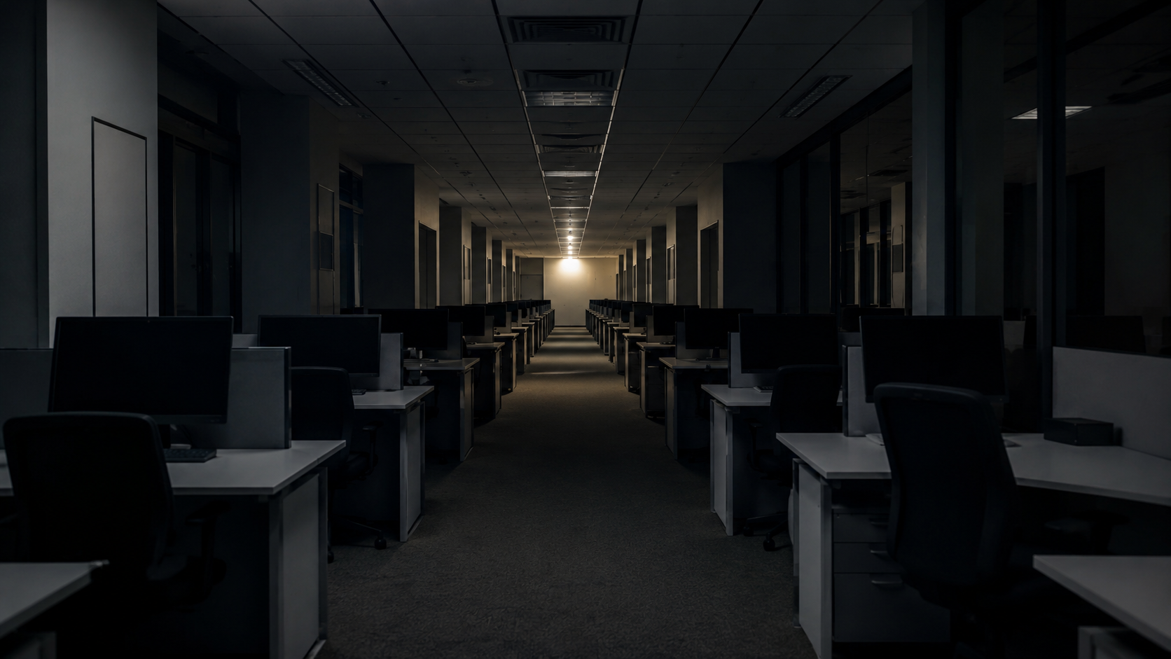 Long alignement de bureaux vides aux écrans éteints, un seul plafonnier allumé au fond du couloir