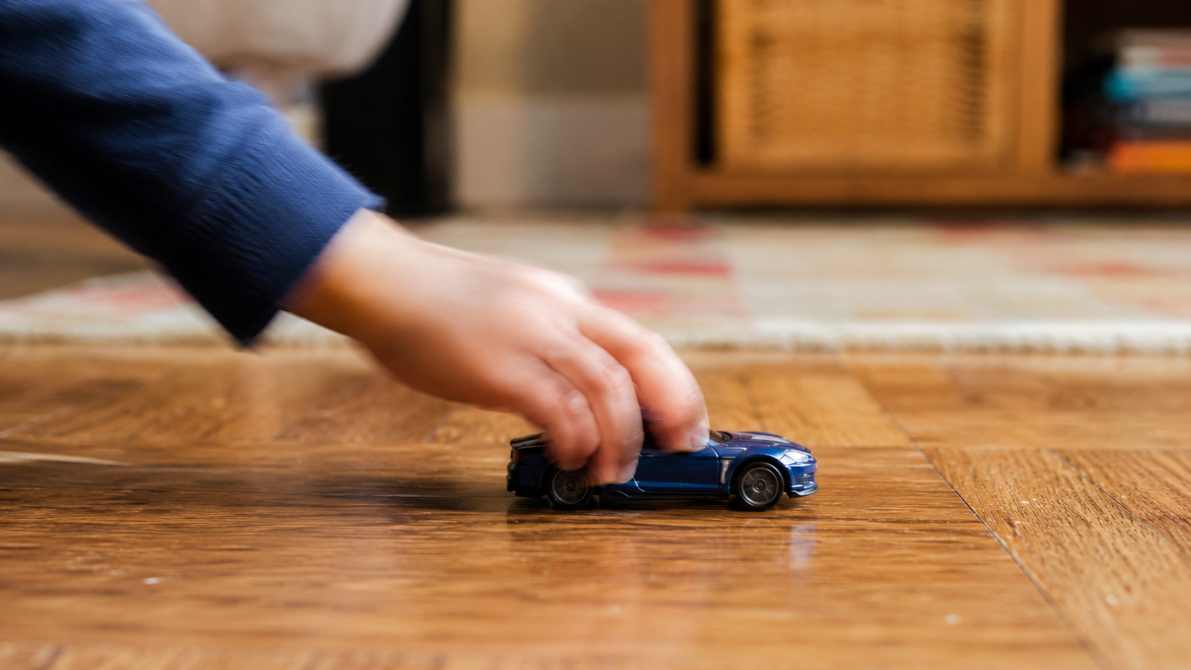 Une main d'enfant floue recule une petite voiture jouet sur un parquet, sans visage visible