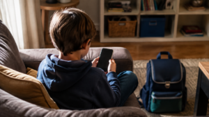 Enfant vu de dos assis sur un canapé, un smartphone à la main, cartable posé au sol.