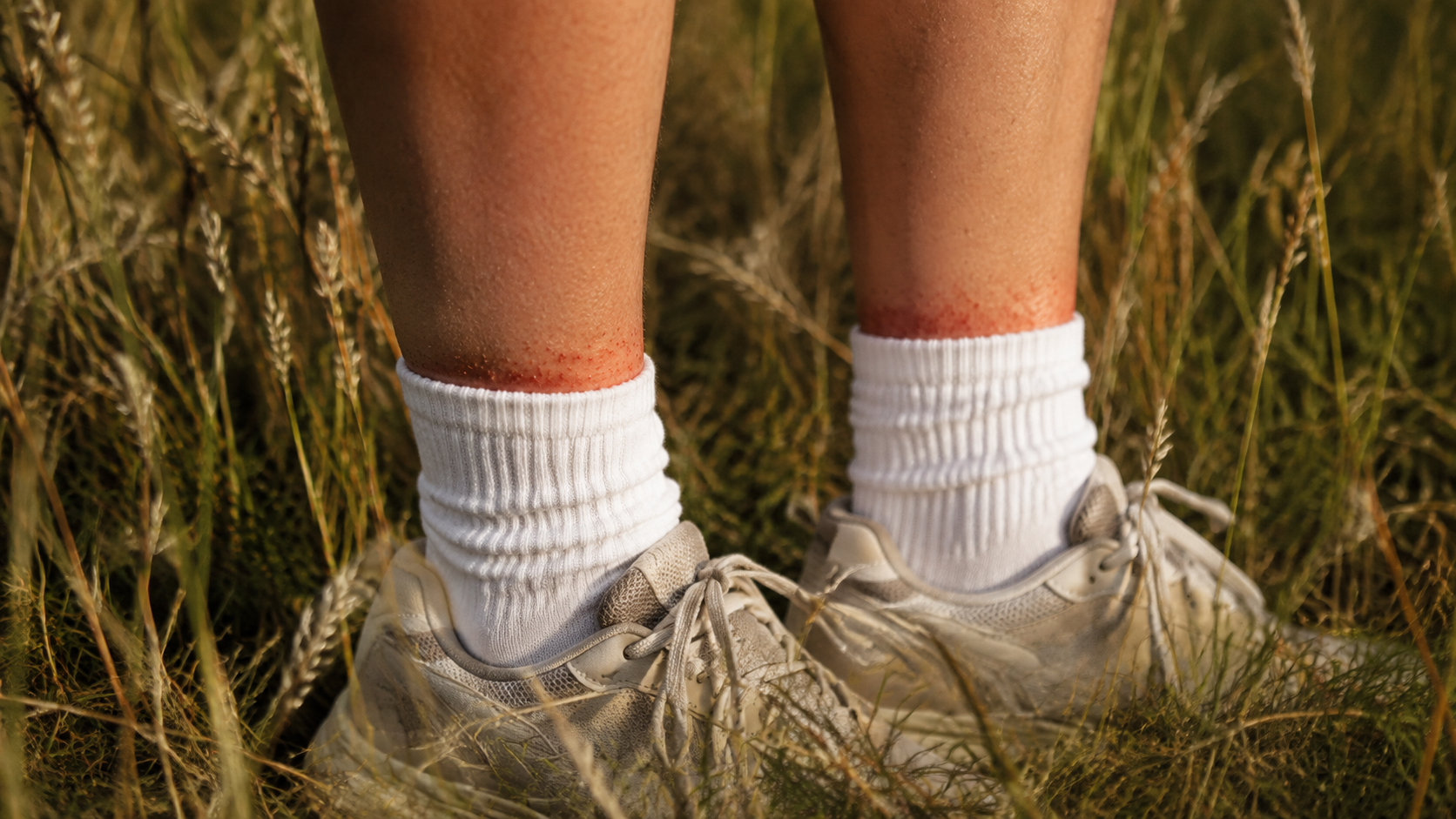 Chevilles en chaussettes blanches et baskets, cadrées sous le genou, au milieu de l'herbe haute.