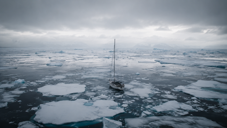 Un voilier de plaisance seul sur une mer arctique grise, entouré de plaques de glace flottantes sous un ciel bas.
