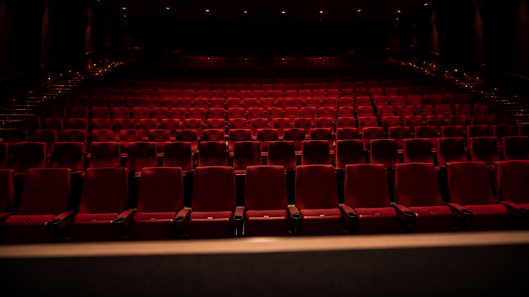 Une salle de cinéma vide photographiée depuis le bord de l’écran, les rangées de fauteuils rouges descendant dans la pénombre.