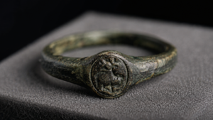 Petite bague-sceau ancienne en métal sombre et corrodé, posée sur une mousse grise de conservation sous une lumière rasante.