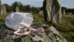 Petit bonnet de baptême blanc en broderie anglaise, noué d’un ruban rose pâle, posé sur la pierre grise d’un menhir.