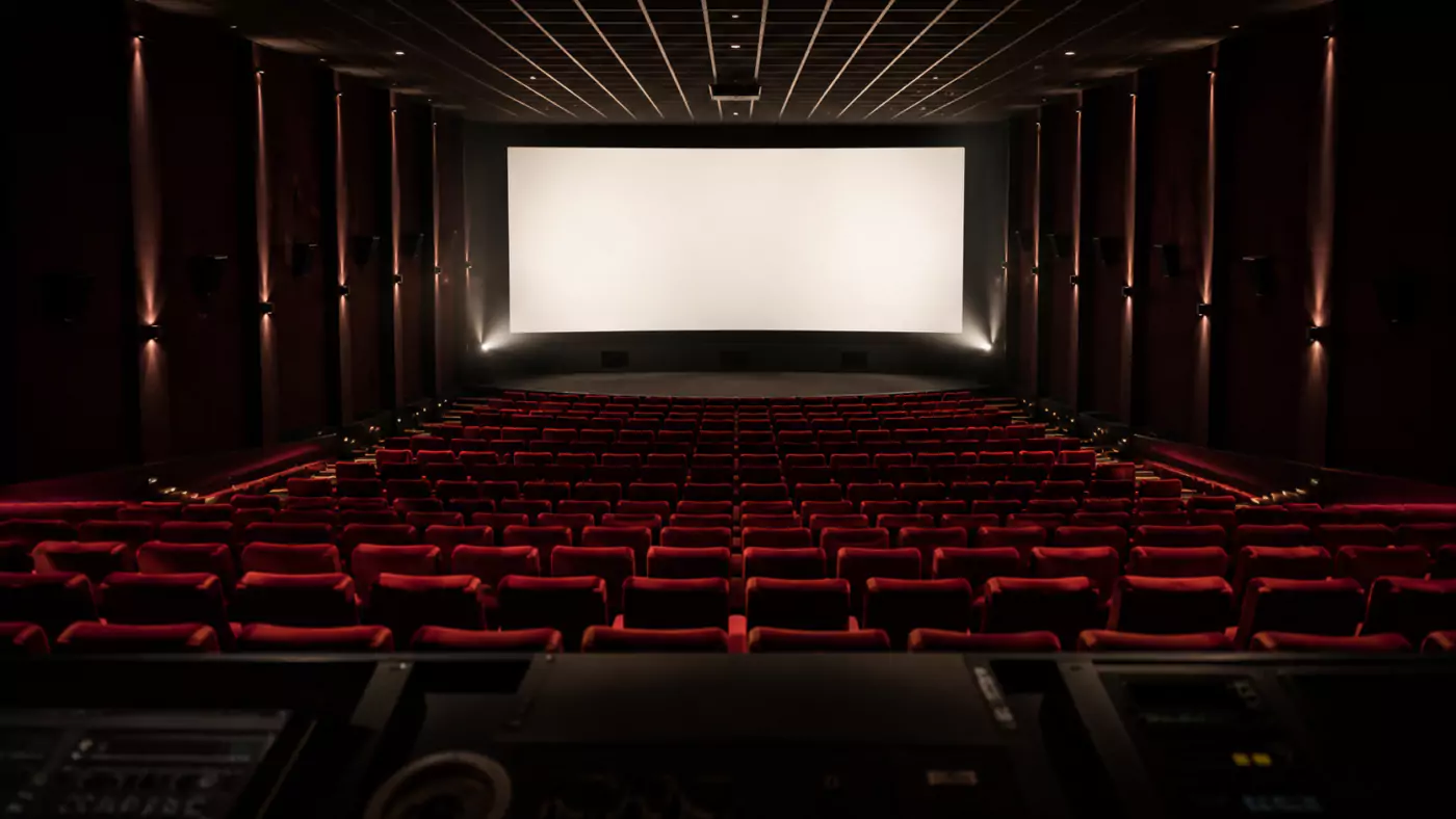 Salle de cinéma vide vue depuis la cabine de projection, fauteuils rouges alignés et écran blanc allumé