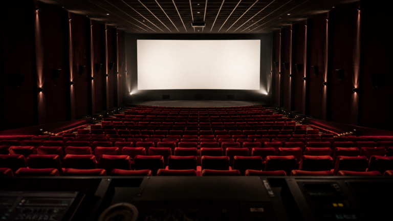 Salle de cinéma vide vue depuis la cabine de projection, fauteuils rouges alignés et écran blanc allumé