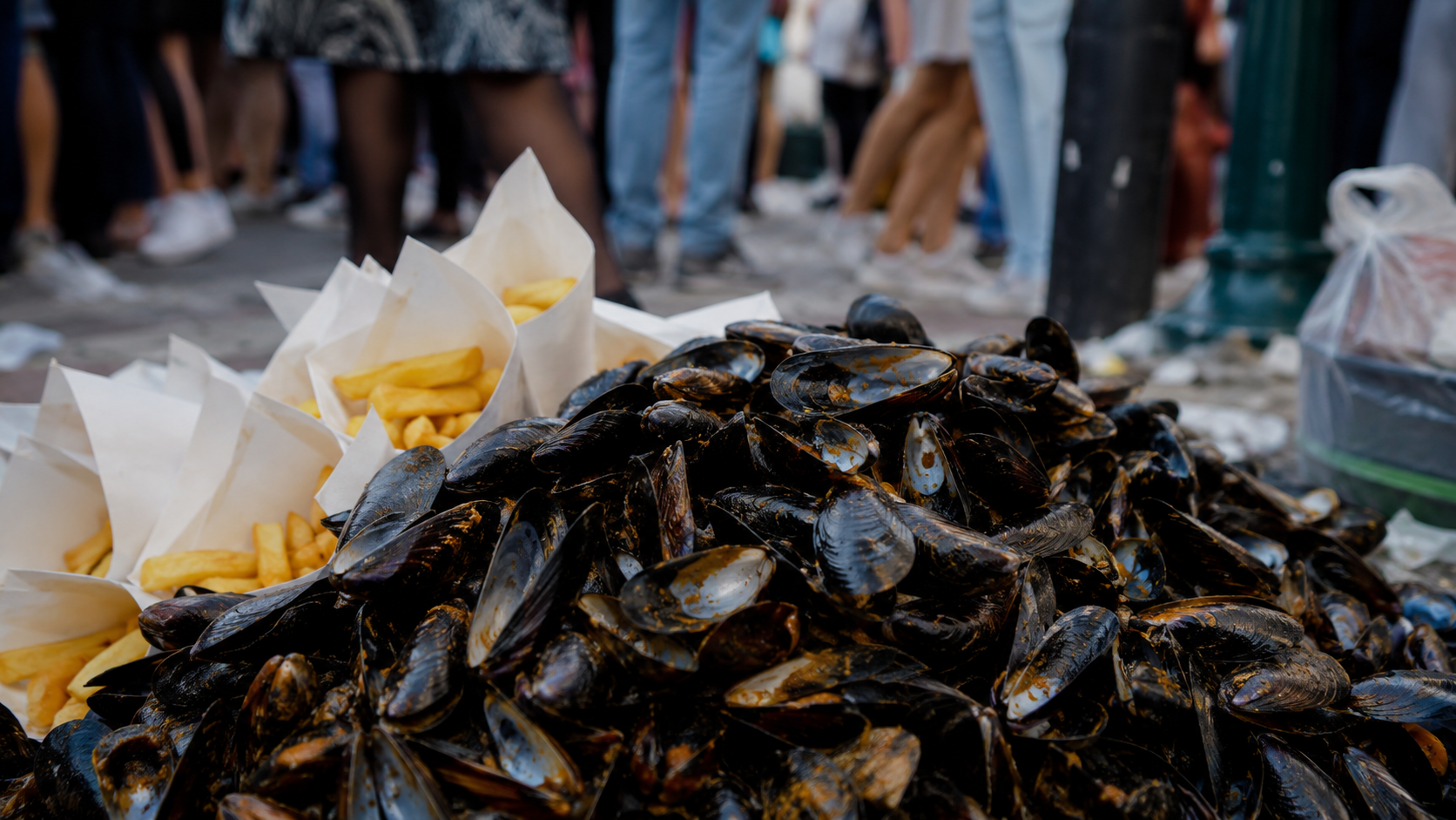 Monticule de coquilles de moules vides amassées sur un trottoir pendant la braderie, cornets de frites à côté.