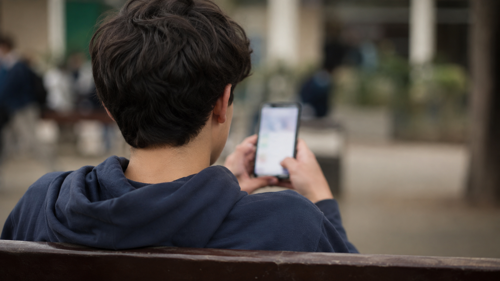 Gros plan sur les mains d’un adolescent tenant un téléphone qui affiche un écran de résultats flou, assis seul sur un banc.