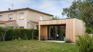 Un studio de jardin en bois clair au fond du jardin d’un pavillon français à enduit beige, toit de tuiles plates et volets roulants, grillage vert et haie en limite de propriété