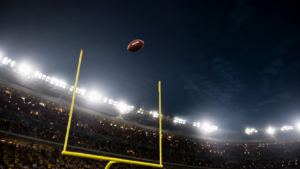 Ballon de football américain au-dessus des poteaux jaunes, filant vers les premiers rangs d’un stade américain éclairé le soir.